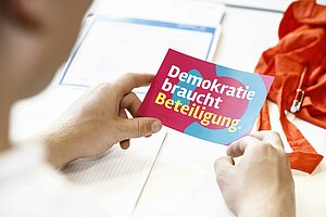 Zwei Hände halten einen Sticker, auf dem "Demokratei braucht Beteiligung" steht