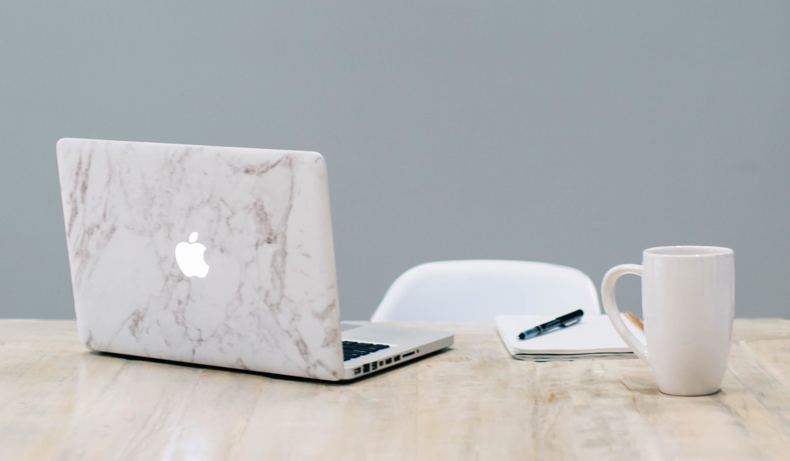 Apple Macbook, ein Notizblock und eine Tasse stehen auf einem Tisch, alle Elemnte sind in hellen Tönen gehalten